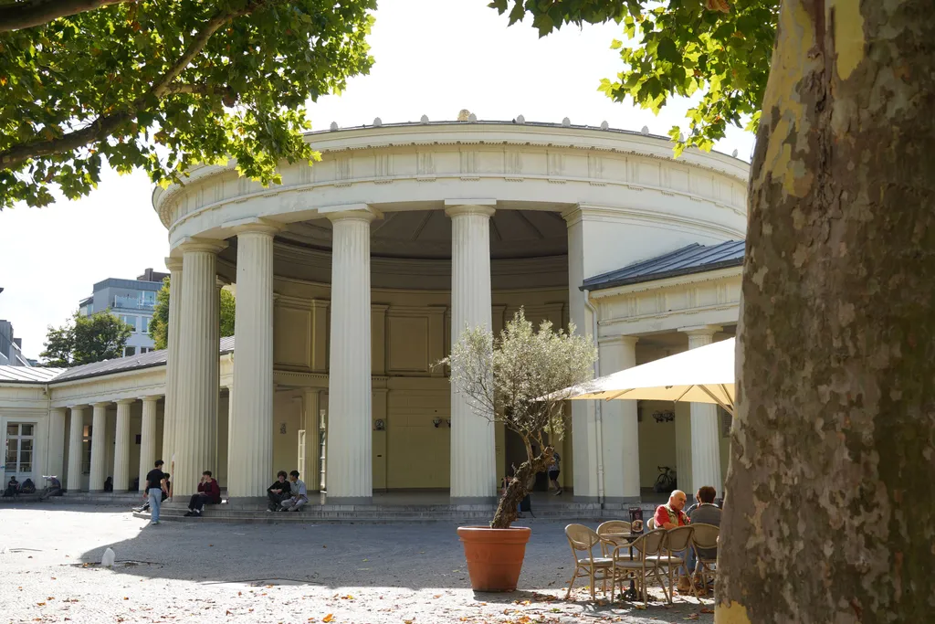 Der Elisenbrunnen in Aachen, ein klassizistischer Pavillon mit hohen weißen Säulen in einer kreisförmigen Kolonnade. Im Vordergrund sitzen Menschen unter einem Baum im Straßencafé und genießen den sonnigen Tag. Die Szene verbindet historische Architektur mit entspanntem Stadtleben.