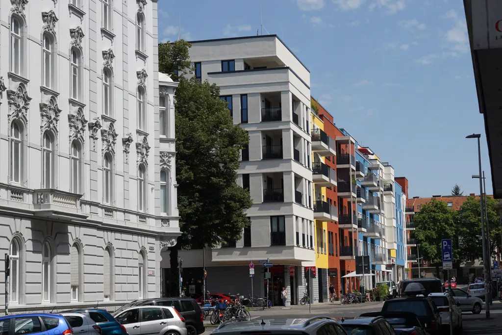 Straßenszene der Viktoriaallee in Aachen mit dem Eckgebäude, in dem cycos ansässig ist. Die Architektur ist modern, mit weißen Fassaden, schwarzen Geländern und großen Fenstern. Die angrenzenden Gebäude sind in kräftigem Blau, Gelb und Rot gestrichen und sorgen für visuelle Vielfalt. Fußgänger und Radfahrer beleben das Straßenbild und vermitteln eine lebendige Nachbarschaftsatmosphäre.