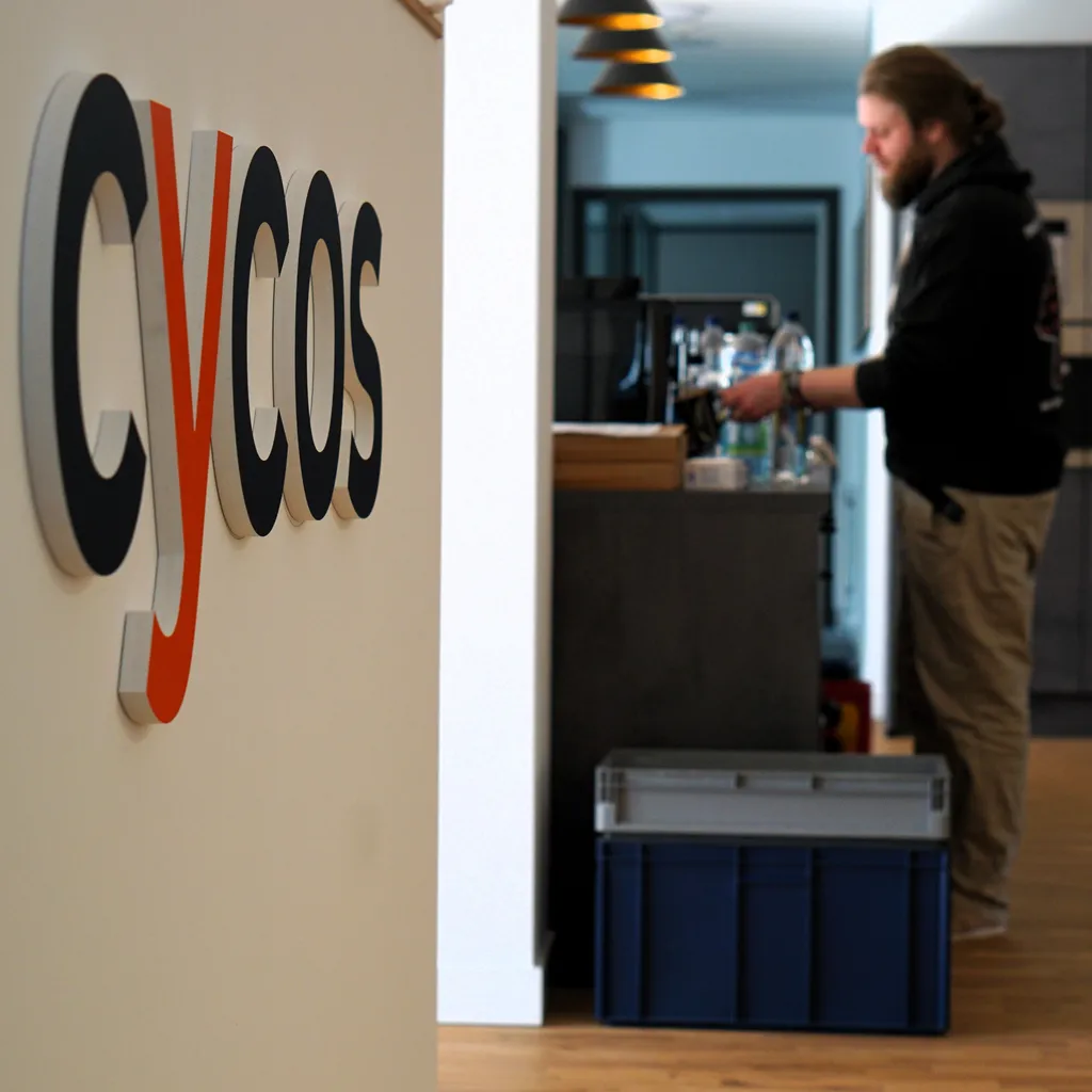 The cycos logo on the wall with someone making coffee in the background