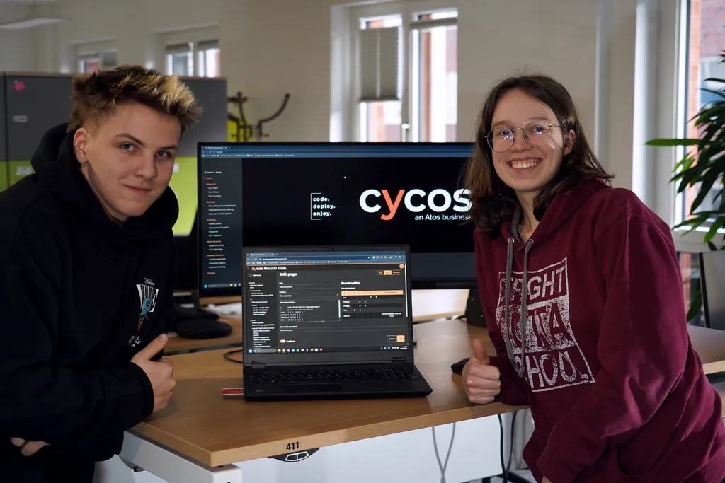 Philipp and Maaike, second‑year trainees at cycos, pose next to a laptop running the NeuralHub CMS editor they developed. Behind them, a large screen shows the cycos branding in a modern office setting.